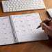 A hand using a pen to write in a Brownline Black DuraFlex 14-Month Planner on a white surface.