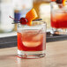 An Acopa rocks glass with a red drink, ice, and fruit garnish on a table in a cocktail bar.