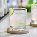 A clear 10 oz. rocks glass filled with a light-colored beverage, ice, a salted rim, and a lime garnish, placed on a round coaster.