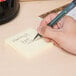 A hand holding a pen and writing on a yellow Universal self-stick note.