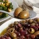 A Vollrath slotted serving spoon with olives being poured from it onto a plate of food.