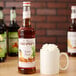 A close-up of a bottle of Monin Butter Pecan flavoring syrup with a white label next to a mug of coffee with whipped cream on top.