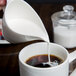 A person pouring white liquid from a white Royal Rideau porcelain creamer into a cup of coffee.