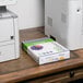 A white rectangular box with a green and white cover of Universal Office Goldenrod Color Copy Paper on a desk next to a Canon printer.