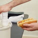 A person using a white Cambro condiment dispenser fixed nozzle pump to add mustard to a hot dog.