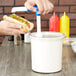 A person holding a hot dog with relish pouring it into a white plastic container with a lid using a black pump.