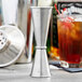 An American Metalcraft stainless steel Japanese style jigger on a table with a drink in the background.