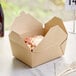 A Kraft paper take-out box with food inside and a wooden fork on a white surface.