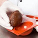 A gloved hand using a Matfer Bourgeat orange silicone half sphere mold to hold a brown round object.