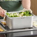 A person holding a Vigor 1/2 size stainless steel pan of broccoli and cauliflower.