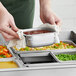 A person holding a Vigor stainless steel hotel pan over a tray of food.