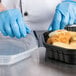 A person wearing blue gloves putting food in a Solo black plastic container.