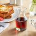 An Acopa glass carafe filled with brown liquid on a table with a glass of liquid, toast, and a white cup.