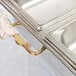 A gloved hand using a Vollrath adapter bar to hold a long stainless steel pan over a white counter.