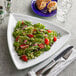 A plate of salad with tomatoes, lettuce, and other vegetables with a fork and knife on a Tuxton Concentrix white triangle china plate.