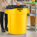 A woman standing next to a yellow Rubbermaid BRUTE trash can.