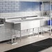 A stainless steel Advance Tabco three compartment sink with two drainboards over a sink with a faucet.