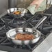 A hand using a pot holder to cook meat in a Vollrath stainless steel fry pan on a stove.