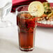 A clear Carlisle plastic tumbler filled with brown liquid and ice with a lemon slice on top.
