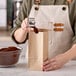 A person using a metal cup to pour coffee into a brown paper bag.