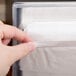 A hand using a Vollrath black countertop napkin dispenser with clear faceplate to take a napkin.