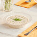 A WNA Comet clear plastic bowl filled with soup on a table with a silver spoon and a glass of water.