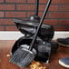 A Rubbermaid upright dustpan with cover on the floor next to a broom.