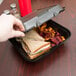 A hand taking a sandwich out of a Genpak black foam container with chips inside.