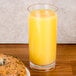 A bagel with cream cheese next to a Cheers clear plastic highball glass filled with orange juice.