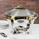 A Vollrath Panacea large round chafing dish with gold and silver accents on a table outdoors.