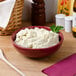 A white Tuxton bowl filled with mashed potatoes on a table with a wooden spoon.