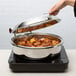 A hand holding a Vollrath stainless steel glass lid over a pot of food.