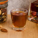 A 1.5 oz. plastic shot glass filled with brown liquid on a table next to a jar of nuts and a bottle of liquor.