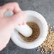 A person's hand grinding brown seeds in a white marble mortar with a white pestle.