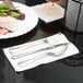 A table set with a WNA Comet linen-like napkin, stainless steel look cutlery, and a plate of food with a fork and spoon.