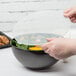 A person putting a plastic lid on a black plastic bowl of salad.