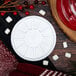 A Dart white foam lid with a circular pattern on a wood table with a red and white plate of marshmallows and candy.