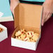 A person opening a 6" x 6" white bakery box of pastries on a table.