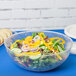 A salad in a clear Fineline plastic bowl on a table with bread and other food.