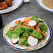 A Fineline white plastic salad plate with blue and gold rim on a table with a salad.