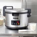 A silver and black Hamilton Beach commercial rice cooker on a table with white rice, a white bowl, and a plastic spoon.