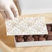 A gloved hand placing a white pad with gold floral designs in a box of chocolates.