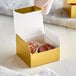 A hand holding a gold foil candy box with a chocolate truffle inside.