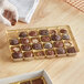 A gloved hand holding a 9 5/16" x 6" x 3/4" gold candy tray with small round chocolates inside.