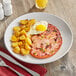 An Acopa bright white stoneware plate with ham and eggs and a side of potatoes and a knife.