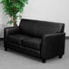 A black leather Flash Furniture loveseat in a lounge area with a plant in front of it.