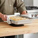 A person holding a Vollrath stainless steel steam table pan with food on a table.