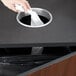 A hand dropping crumpled paper into a Vollrath round stainless steel in-counter trash chute.