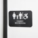 A Tablecraft ADA restroom sign with Braille for a family and wheelchair accessible restroom, in black and white.