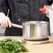 A person pouring a liquid into a Vollrath stainless steel sauce pan with a red lid.